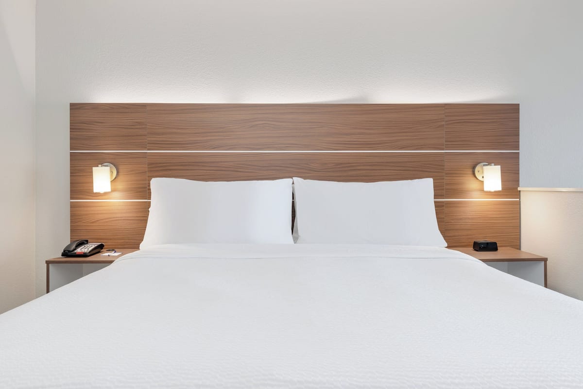 A hotel room with a wooden headboard, two bedside lamps, a white bedspread, and a nightstand with a phone and a remote control.
