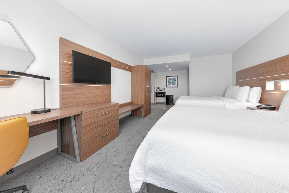 A modern hotel room with a large bed, a desk with a chair, a television, and a small kitchenette area.