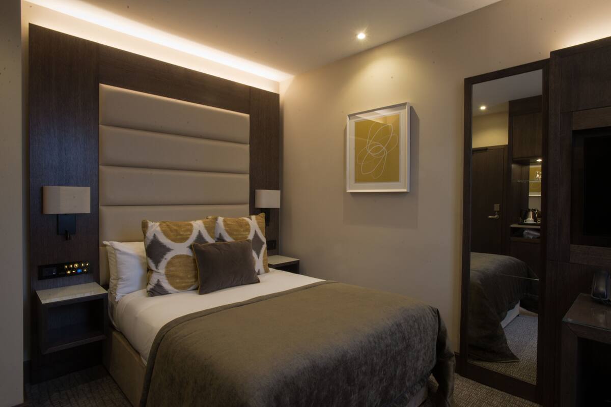 A hotel room with a large bed, a nightstand with a lamp, a wall-mounted artwork, and a mirror.