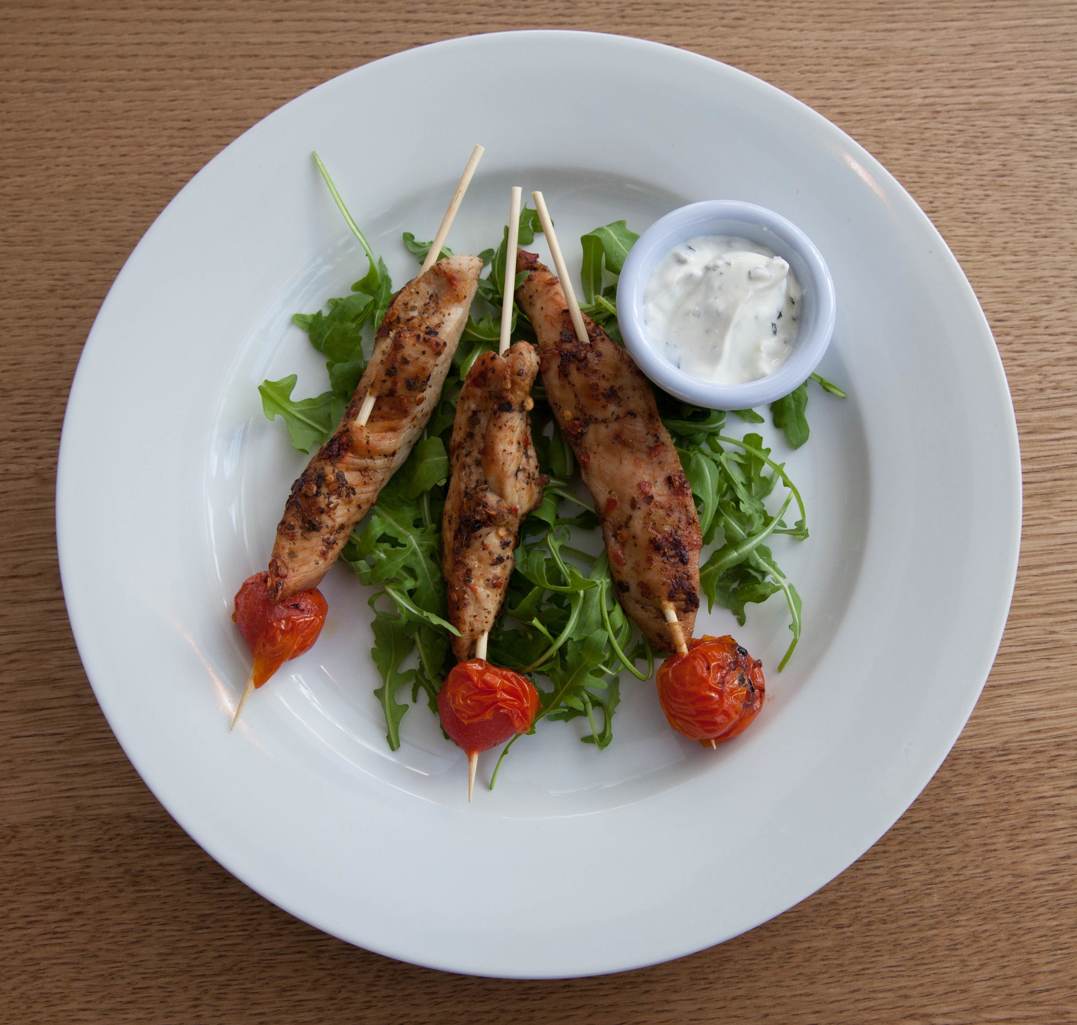 A plate with grilled chicken skewers, cherry tomatoes, and a side of creamy sauce on a bed of arugula.