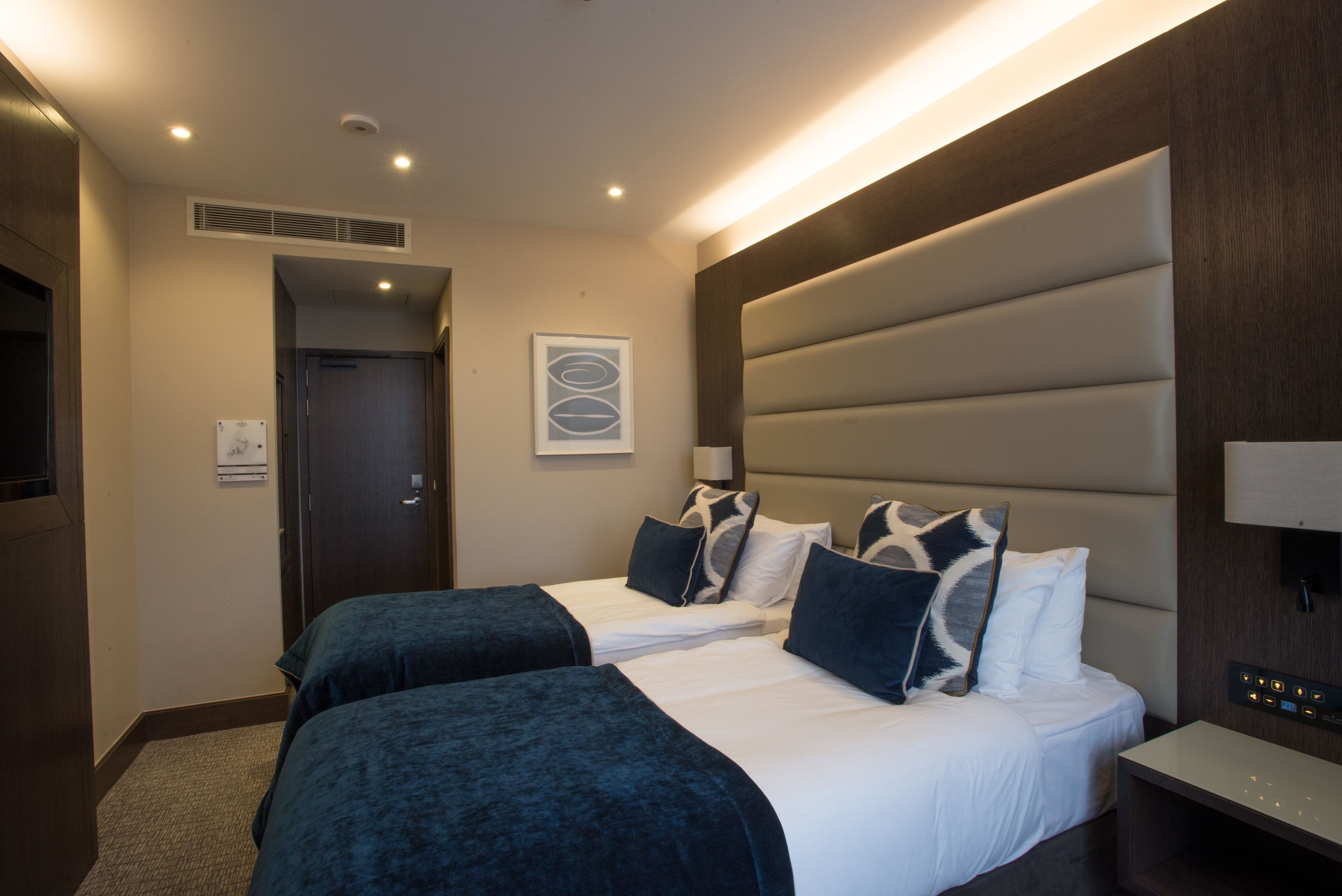 A hotel room with two beds, a large padded headboard, a bedside table, and a wall-mounted TV.