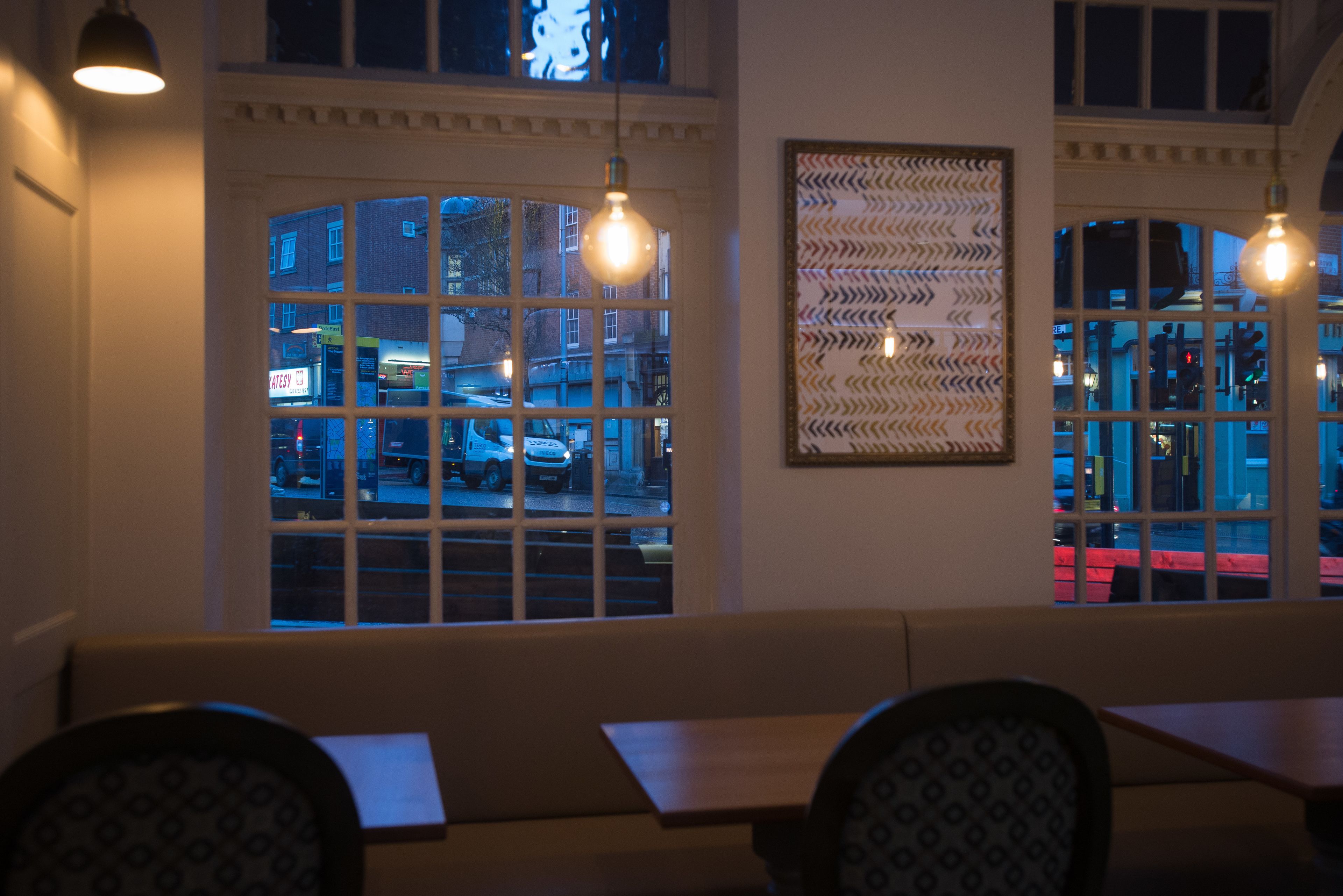 L’intérieur d’un restaurant, avec vue sur la rue grâce à de grandes fenêtres, un mur décoré de motifs et un espace détente avec des bancs.