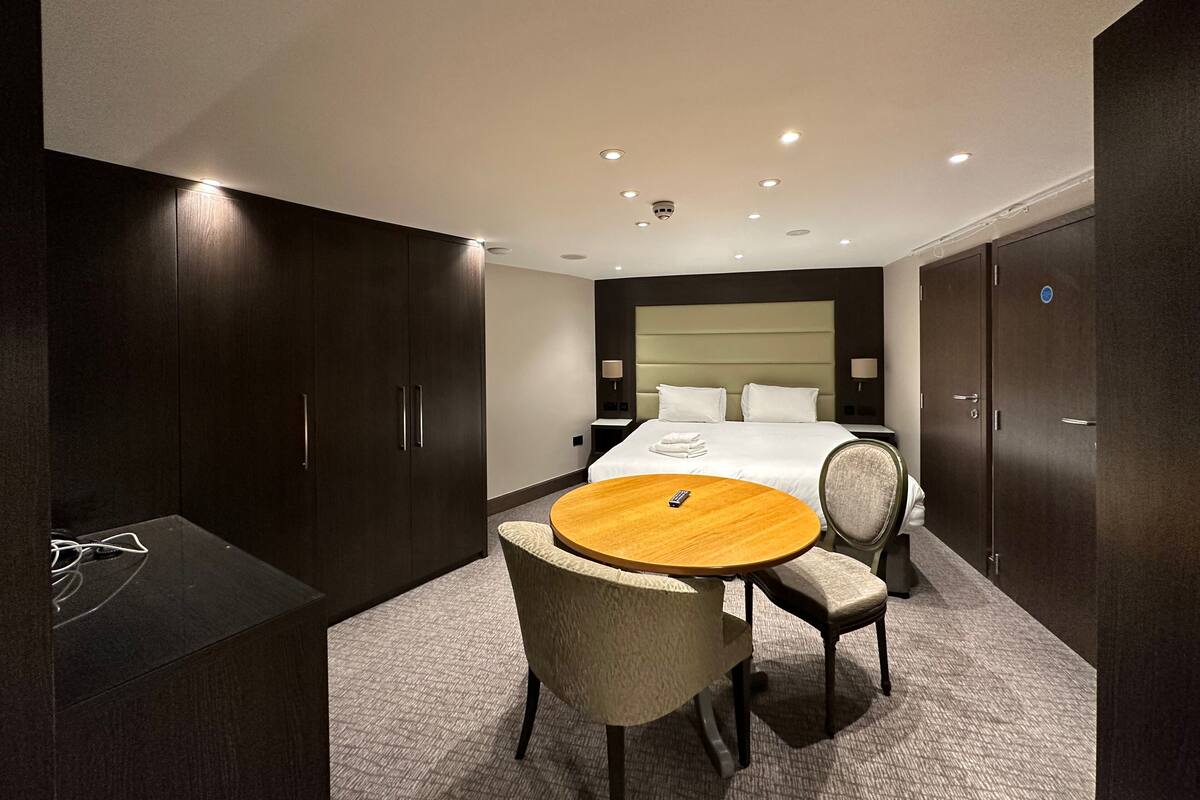 A hotel room with a large bed, a round wooden table, two chairs, a desk with a chair, and a wardrobe.