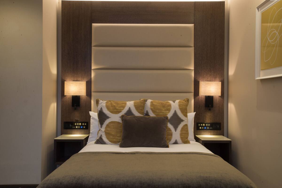 A neatly made bed with a patterned pillow, two bedside tables with lamps, and a wall-mounted control panel.