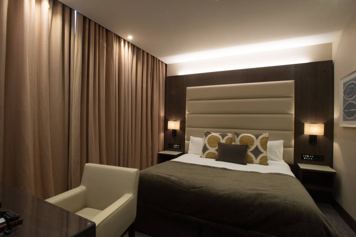 A hotel room with a large bed, a chair, a desk, and a nightstand with a lamp.