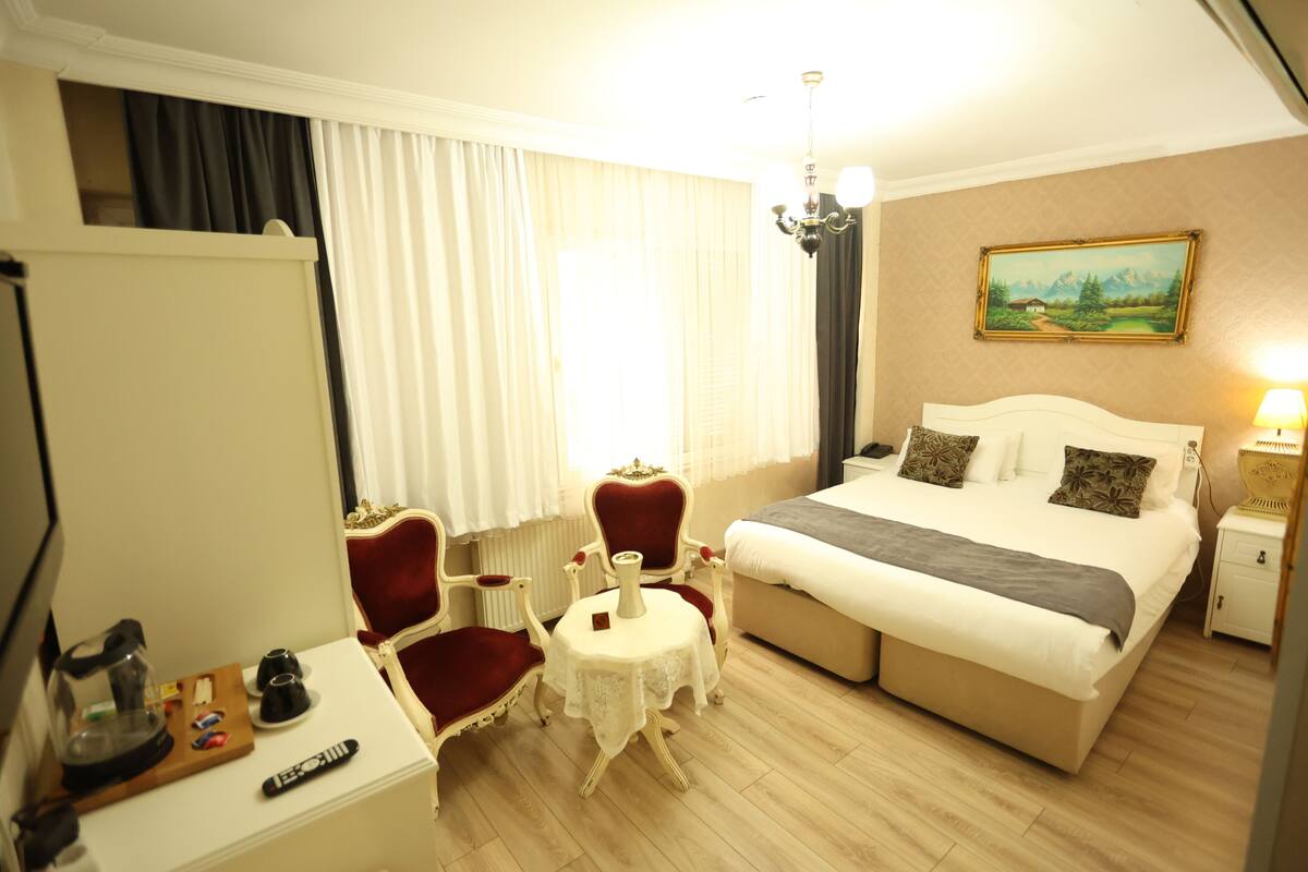 A hotel room with a bed, a desk with a chair, a small table, and a painting on the wall.