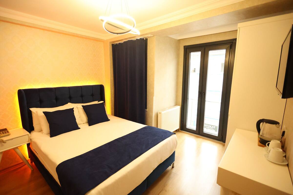 A hotel room with a large bed, a bedside table with a lamp, a window with curtains, and a view of the outside.