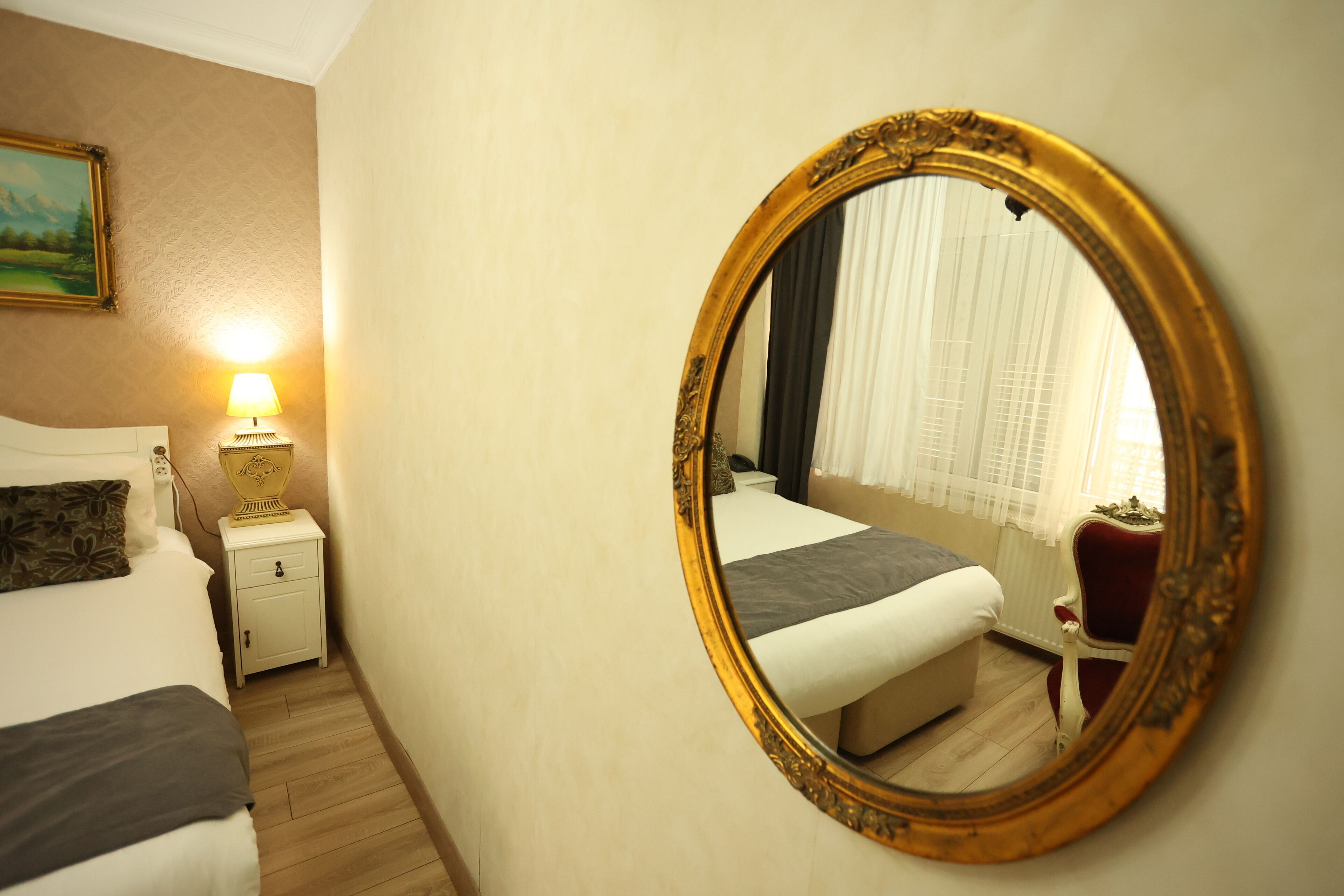 A hotel room with a bed, bedside table with a lamp, a mirror, and a window with curtains.