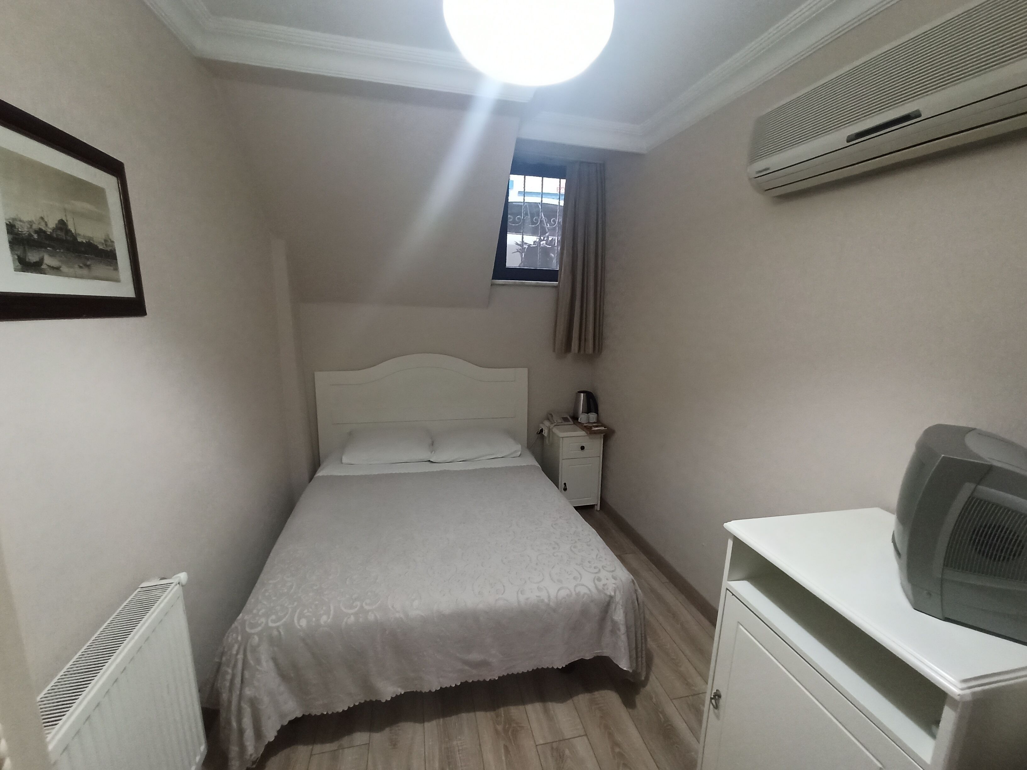 Een kleine, verzorgde slaapkamer met een enkel bed, een nachtkastje met lamp, een radiator en een raam met gordijnen.