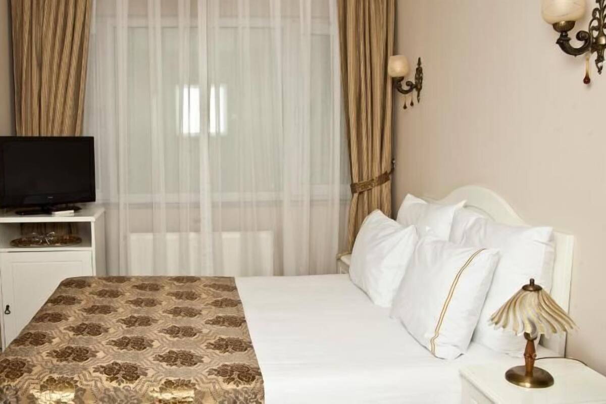 A hotel room with a bed, a TV on a stand, a bedside table with a lamp, and sheer curtains.