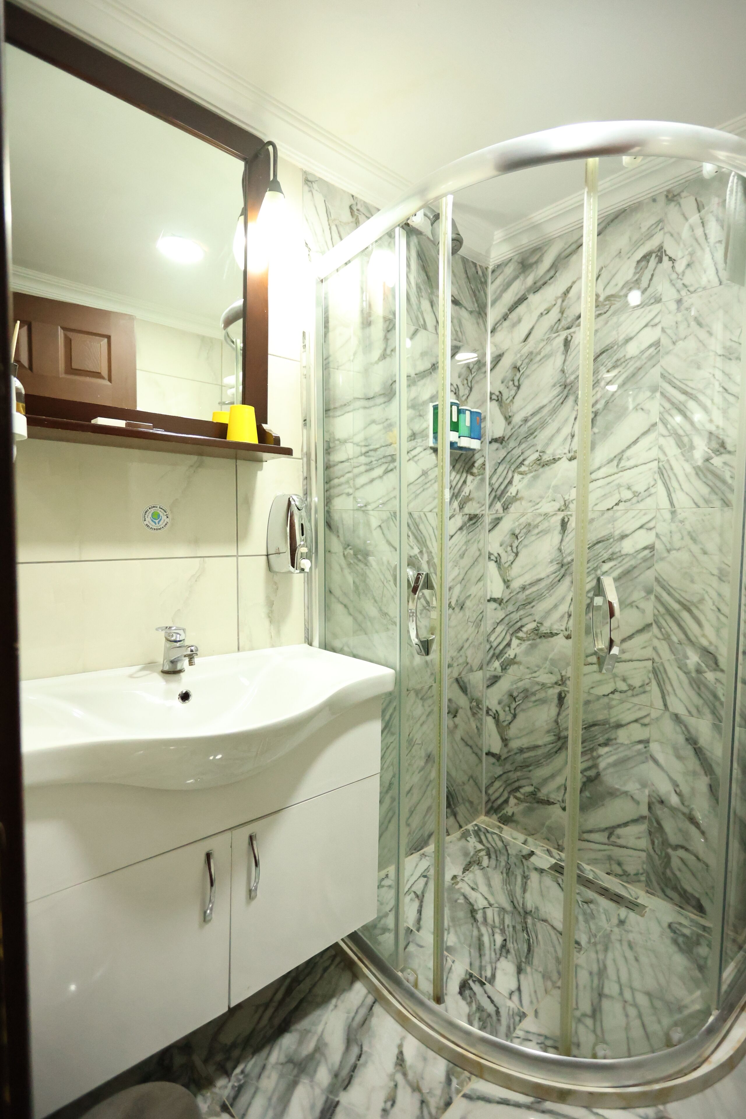 Un baño con pared de mármol, cabina de ducha de vidrio, lavamanos blanco y un espejo.