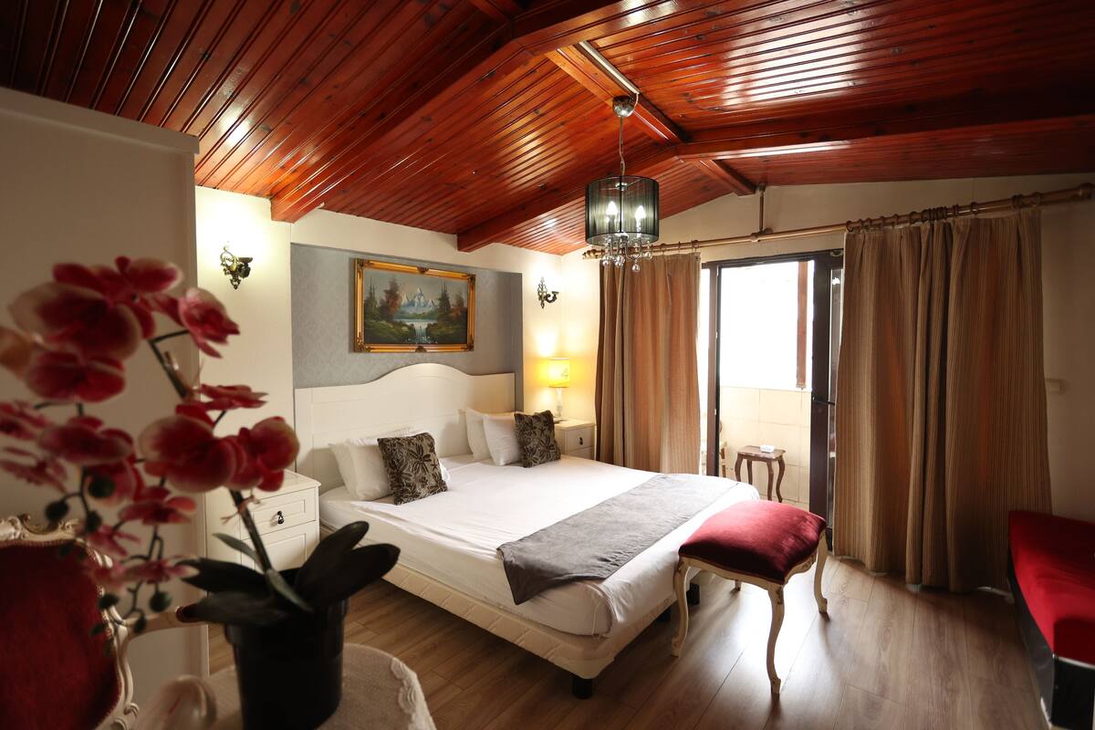 A hotel room with a wooden ceiling, a large bed with a white headboard, a bedside table with a lamp, a red chair, and a window with curtains.