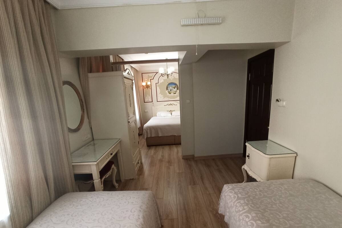 A hotel room with a wooden floor, a desk with a mirror, two single beds, a wardrobe, and a door leading to another room.
