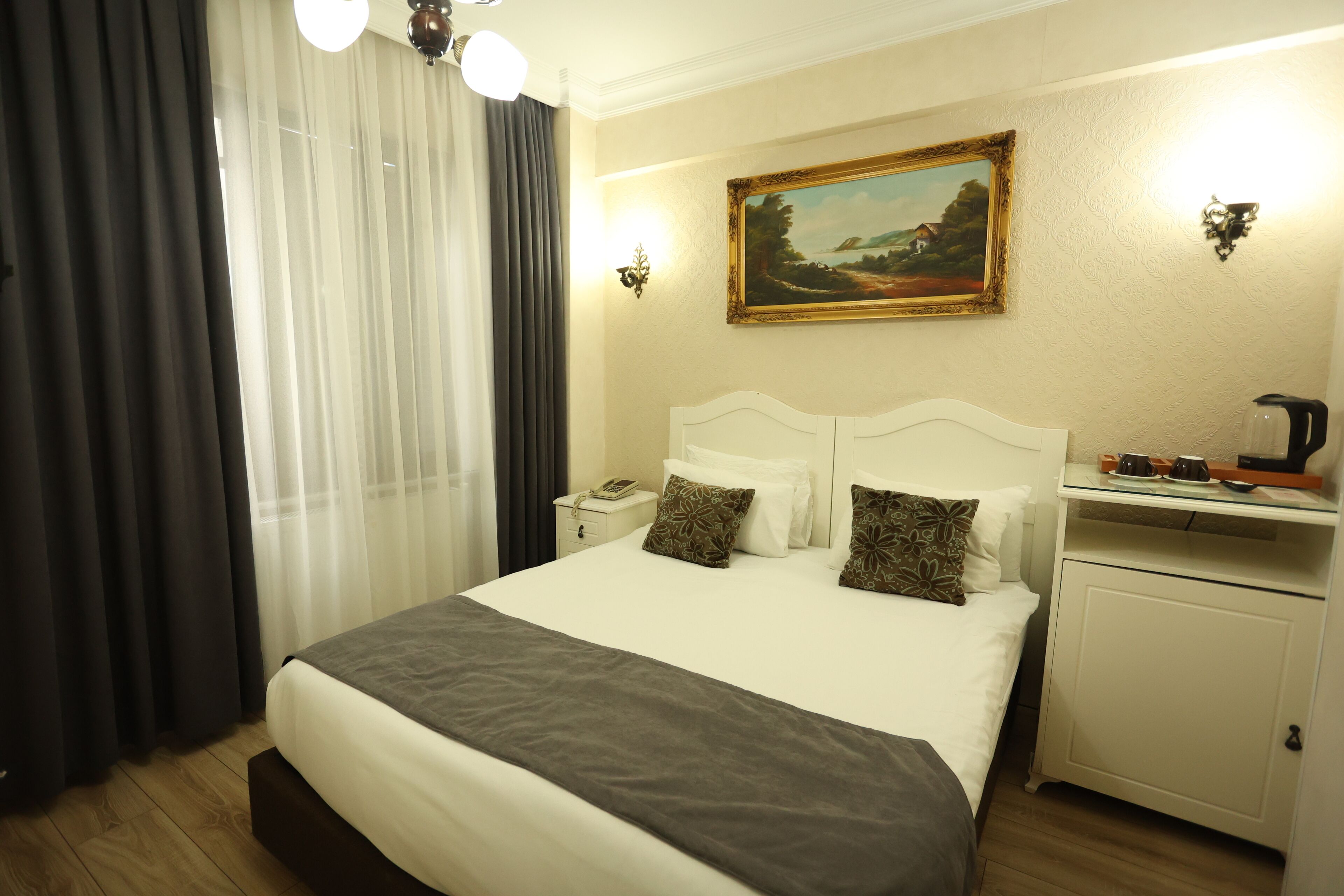 A hotel room with a large bed, a nightstand, a bedside table, a painting on the wall, and a window with curtains.