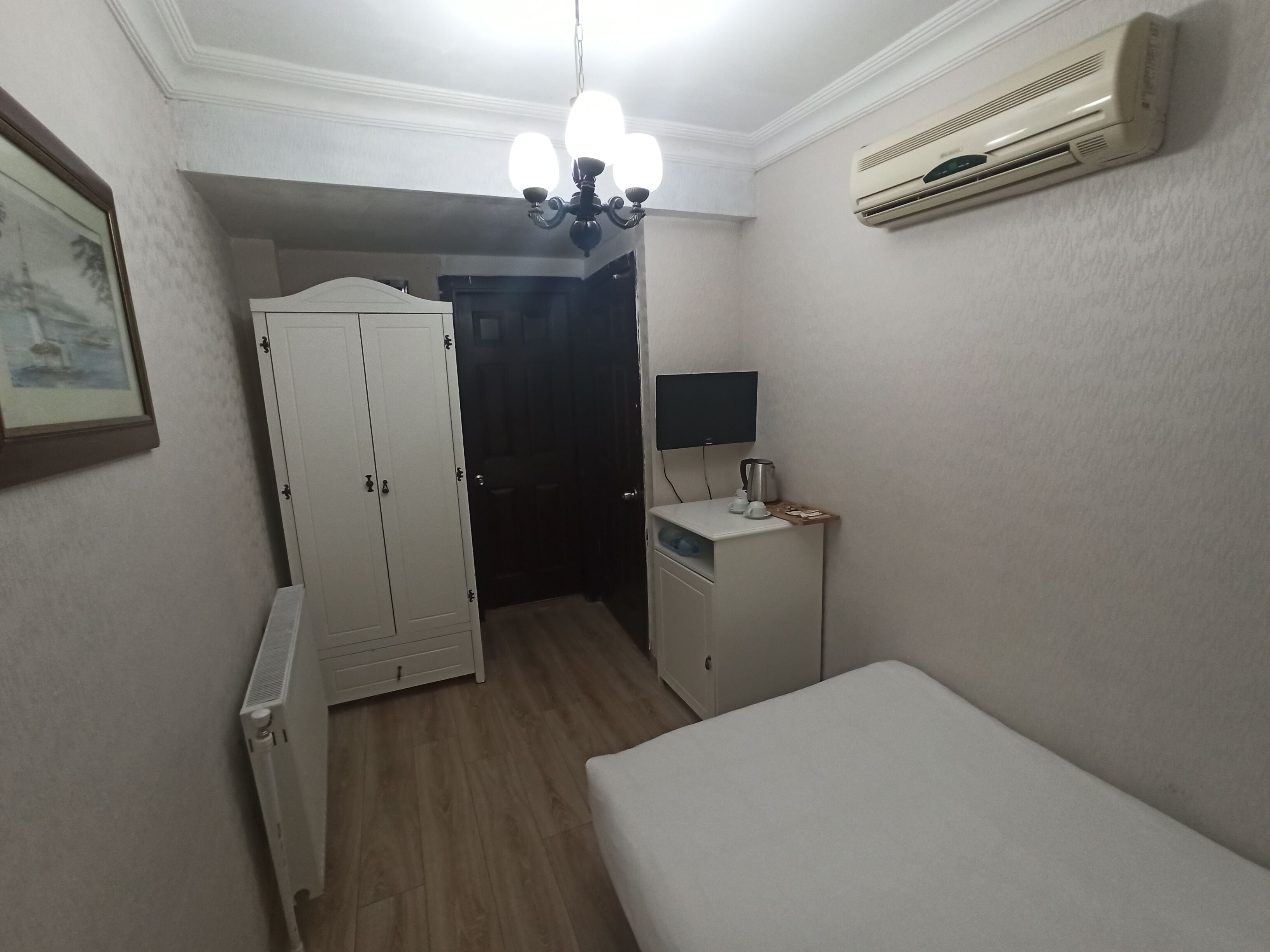 A small, well-lit bedroom with a bed, a radiator, a wardrobe, a desk with a lamp, and an air conditioner.