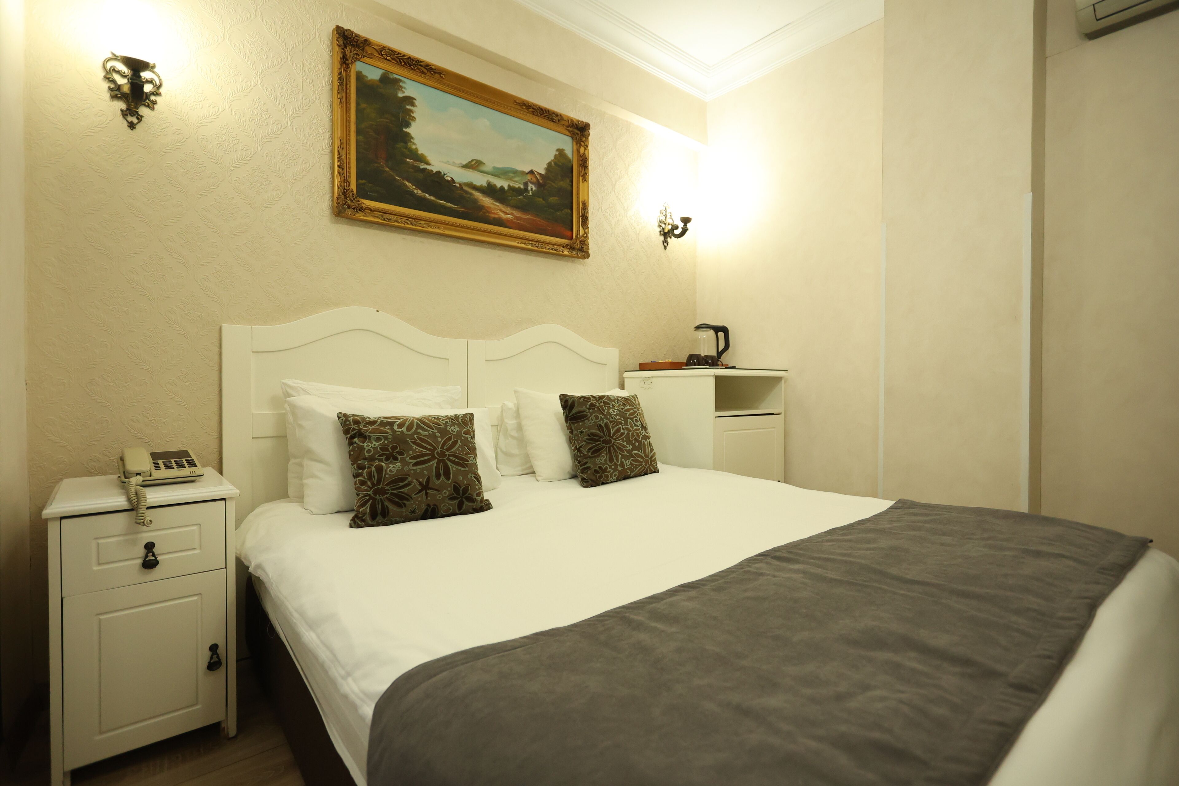 A neatly made bed with a white headboard and two decorative pillows, a bedside table with a telephone, and a painting on the wall.