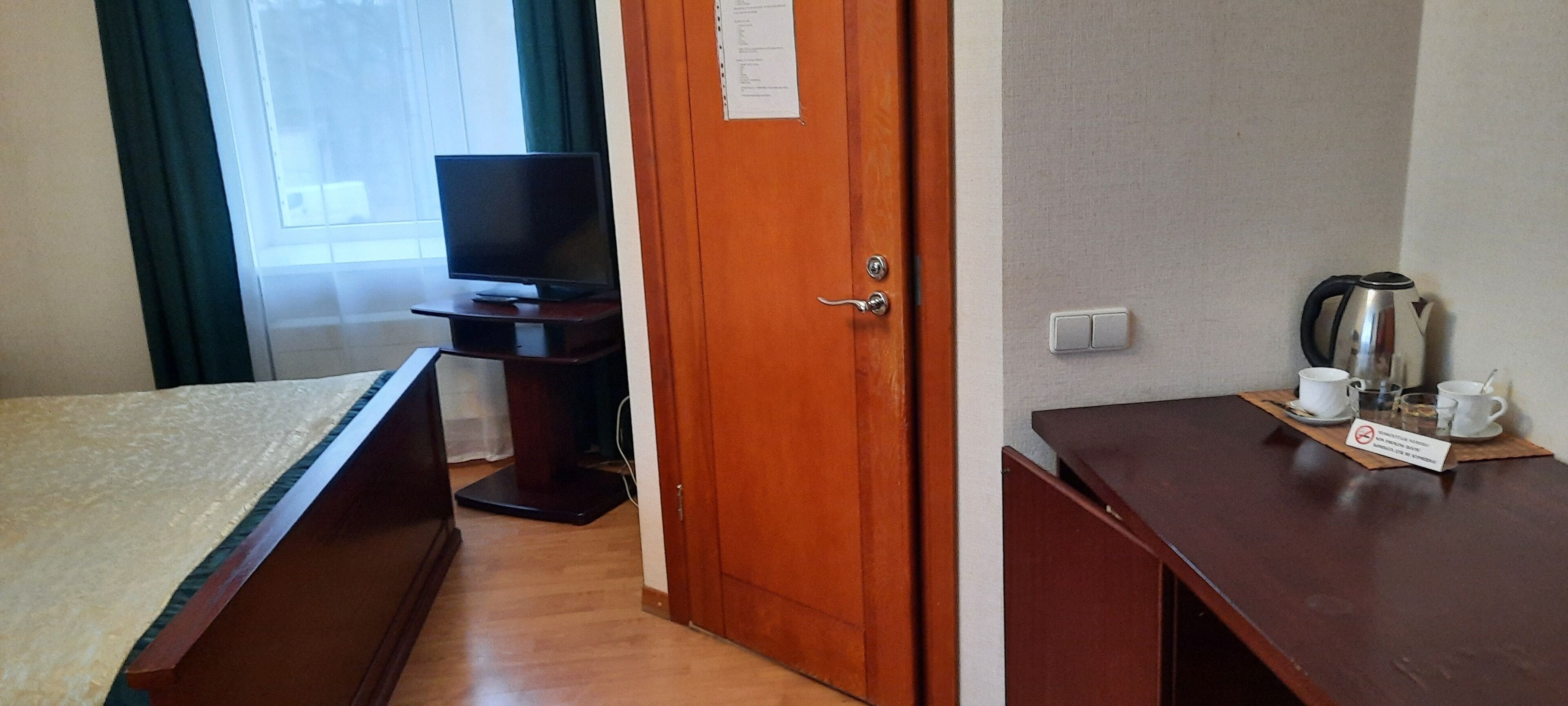 A hotel room with a bed, a TV, a wooden desk with a kettle and cups, and a door with a sign.