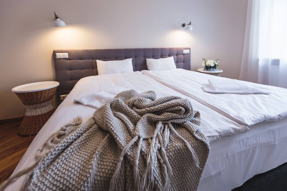 Ein ordentlich bezogenes Bett mit weißen Leinen, einer grauen Strickdecke und einem Nachttisch mit einer Lampe.
