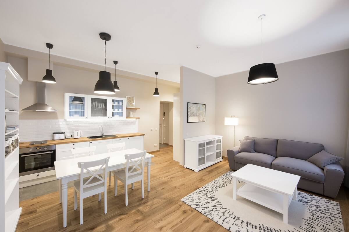 Eine moderne Küche mit weißen Schränken, einem Esstisch und Stühlen. Im Wohnbereich befindet sich ein graues Sofa und ein weißer Couchtisch.