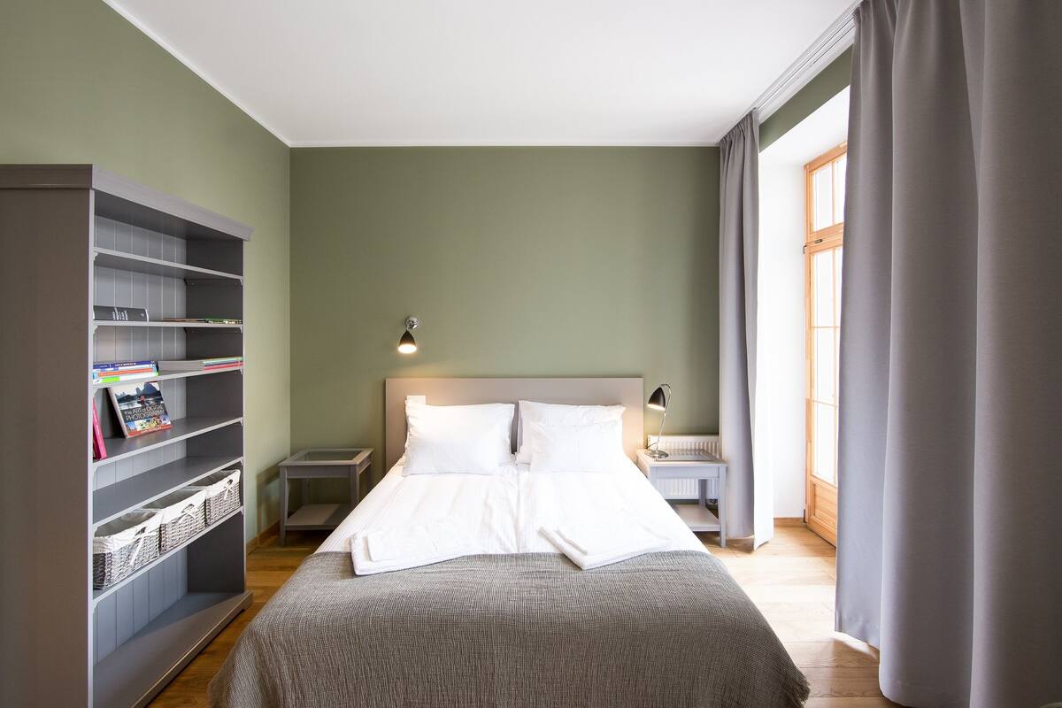 Ein ordentlich bezogenes Bett mit weißen Leinen in einem Zimmer mit grünen Wänden, einem Bücherregal und einem Fenster mit Vorhängen.