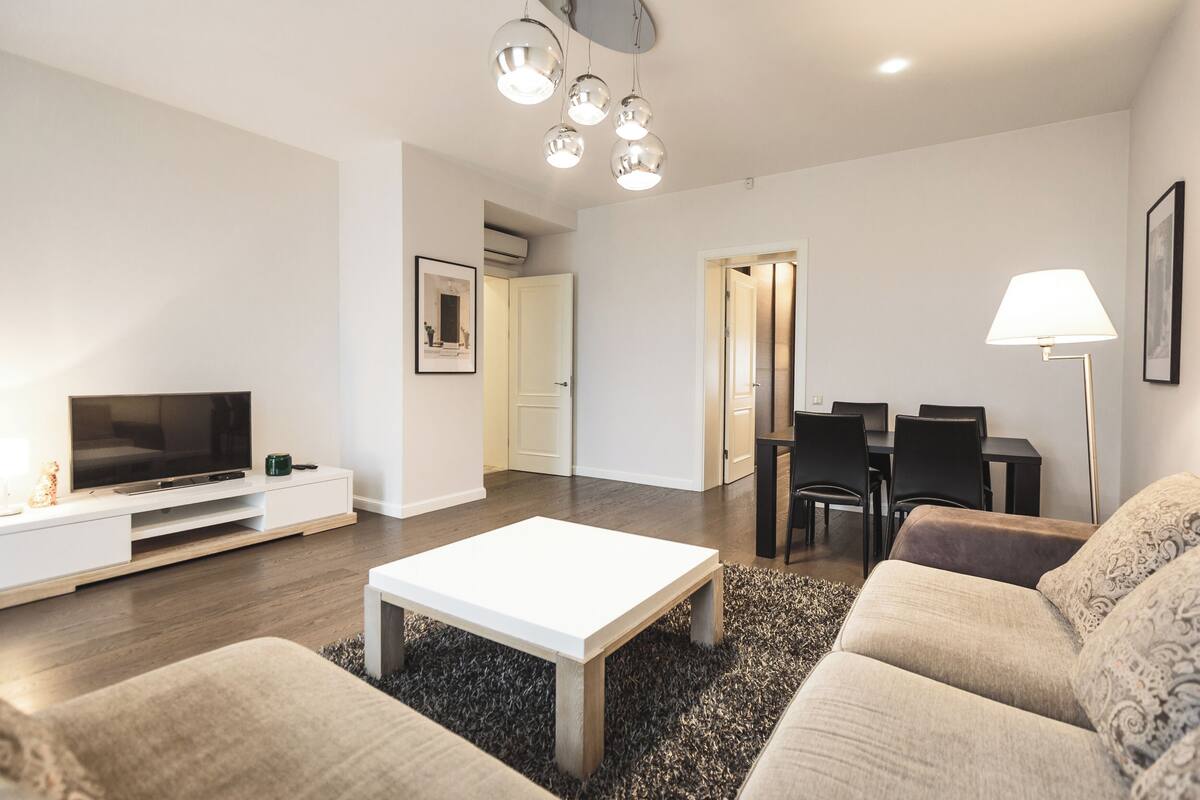 Ein modernes Wohnzimmer mit Flachbildfernseher, weißem Couchtisch, beiger Couchgarnitur und einem Essbereich mit Stühlen und einer Lampe.