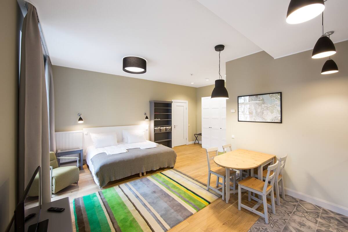 Ein modernes Schlafzimmer mit einem Bett, einem Nachttisch und einem Schreibtisch mit Stuhl. Es gibt einen Esstisch mit Stühlen und einen farbenfrohen Teppich.