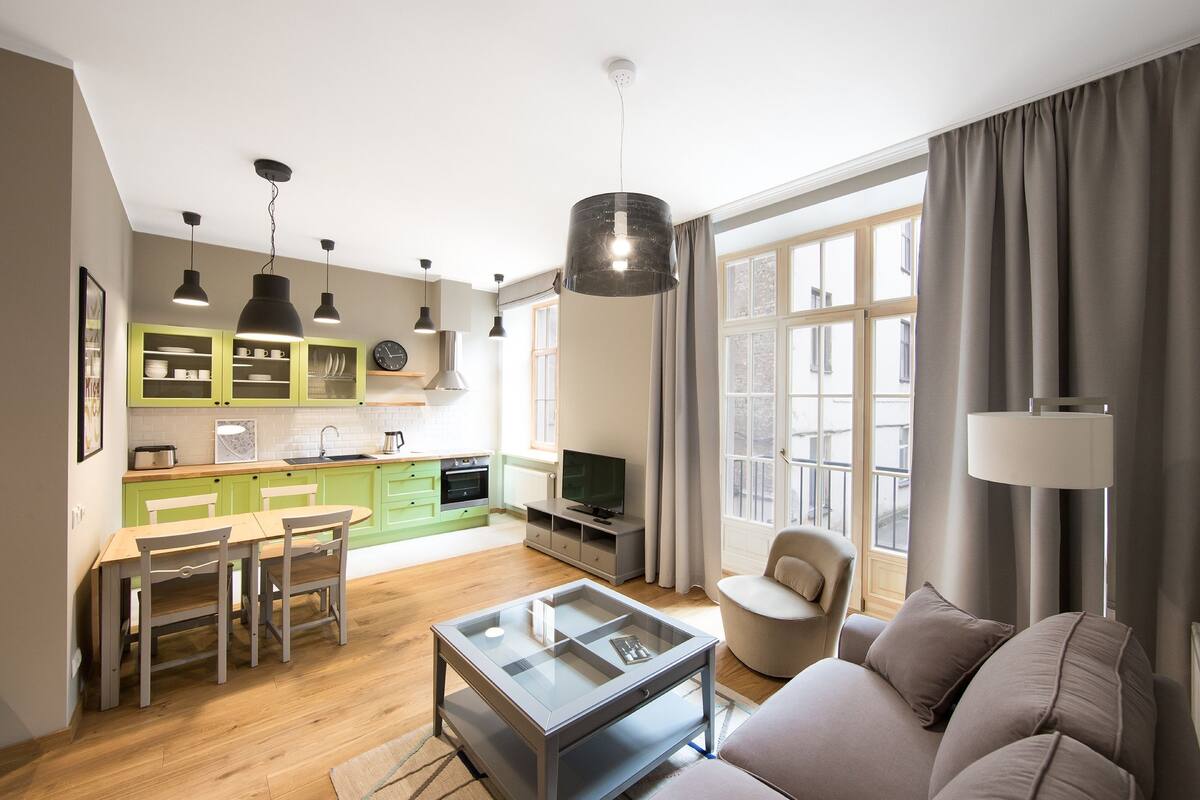 Ein modernes Wohnzimmer mit einer Couch, einem Couchtisch und einem Fernseher. Es gibt einen Essbereich mit Tisch und Stühlen. Das Zimmer verfügt über große Fenster mit Vorhängen und Blick ins Freie.