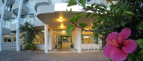 Entrada de hotel com área de espera coberta, escadaria que leva aos andares superiores e uma flor de hibisco rosa em primeiro plano.