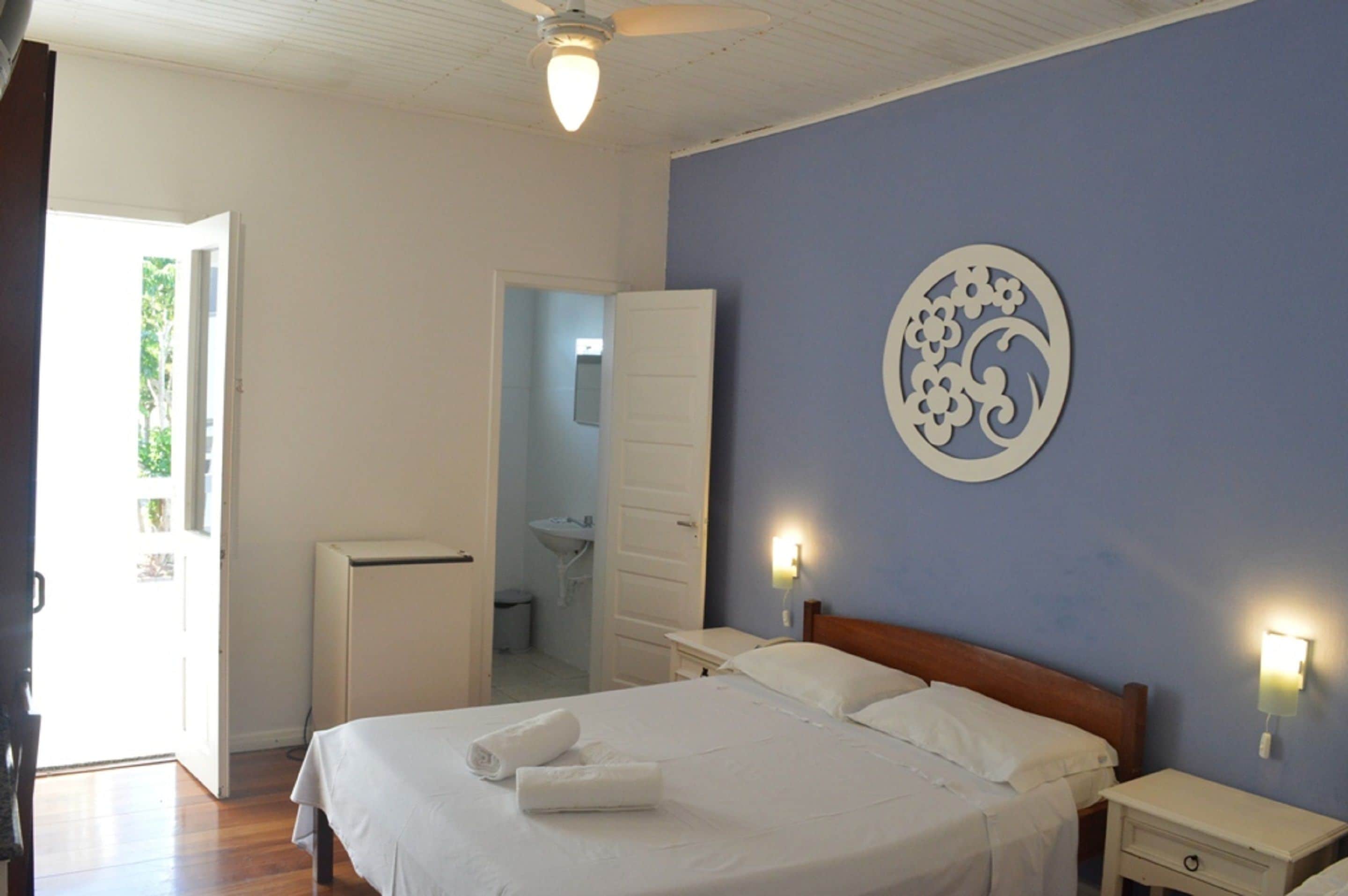 Une chambre à coucher comprenant un lit, des tables de chevet, une lampe fixée au mur et une porte donnant accès à une salle de bain.