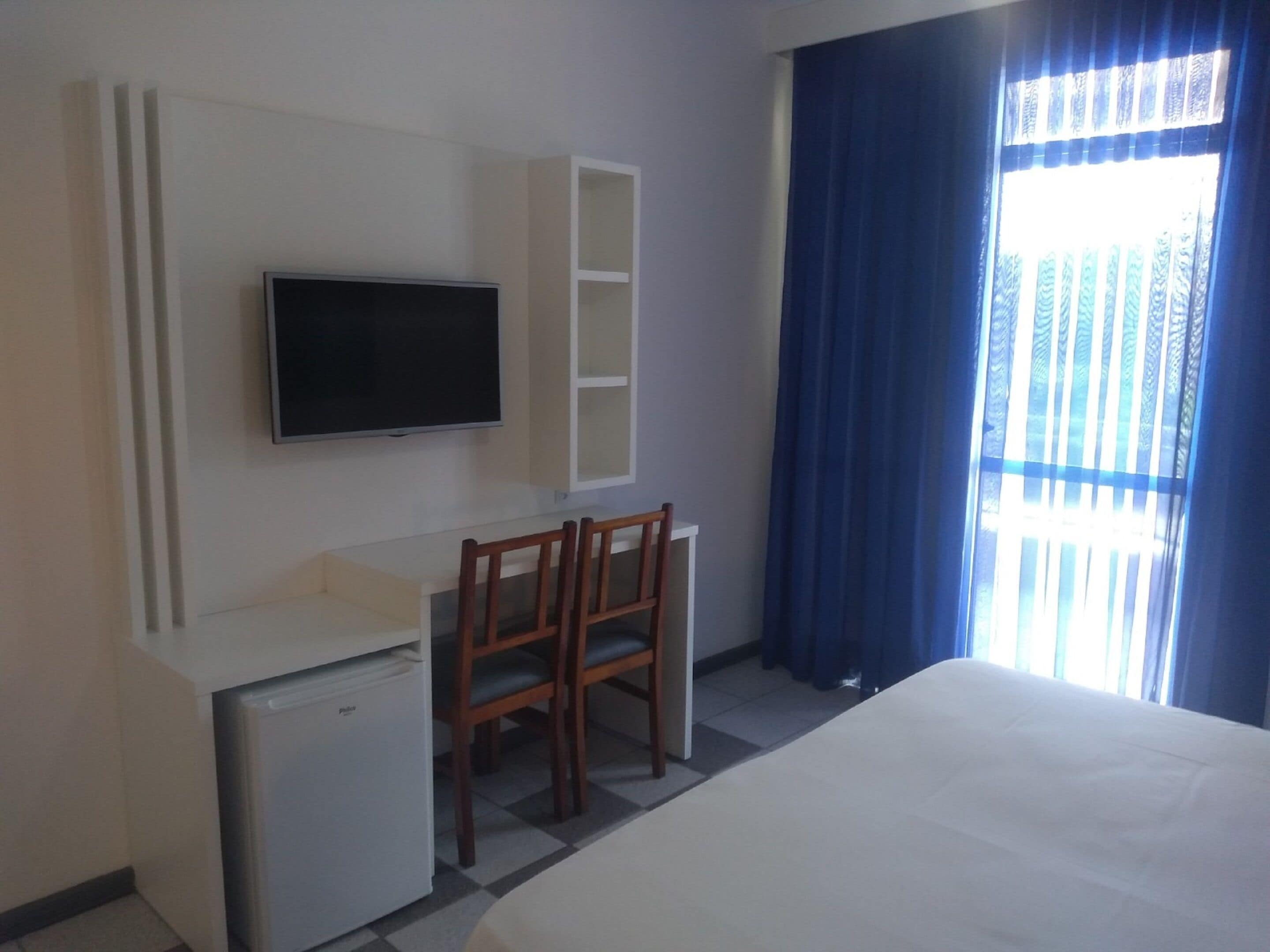 A hotel room with a bed, a desk with a TV, a small fridge, and a window with curtains.