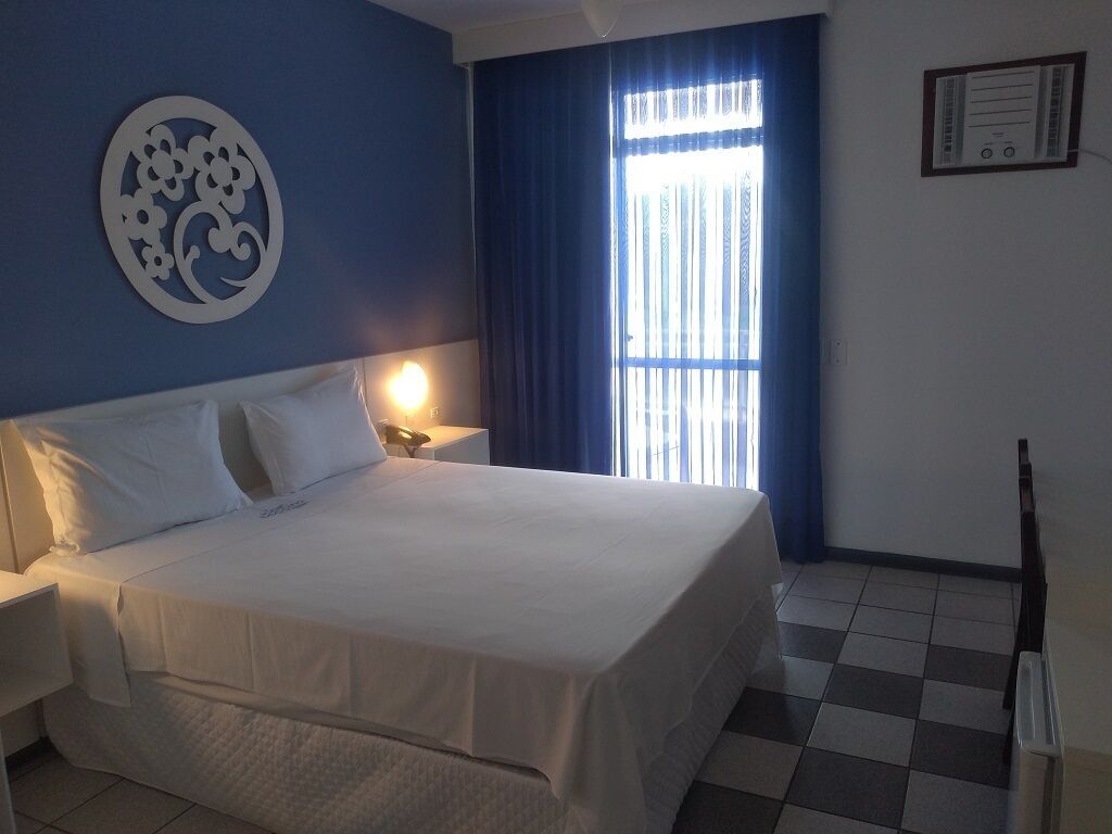 A hotel room with a large bed, a nightstand, a wall clock, and a window with curtains.