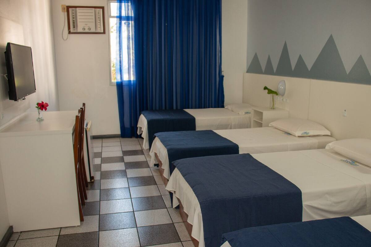 Een hotelkamer met drie eenpersoonsbedden, een bureau met stoel, een televisie en een raam met blauwe gordijnen.