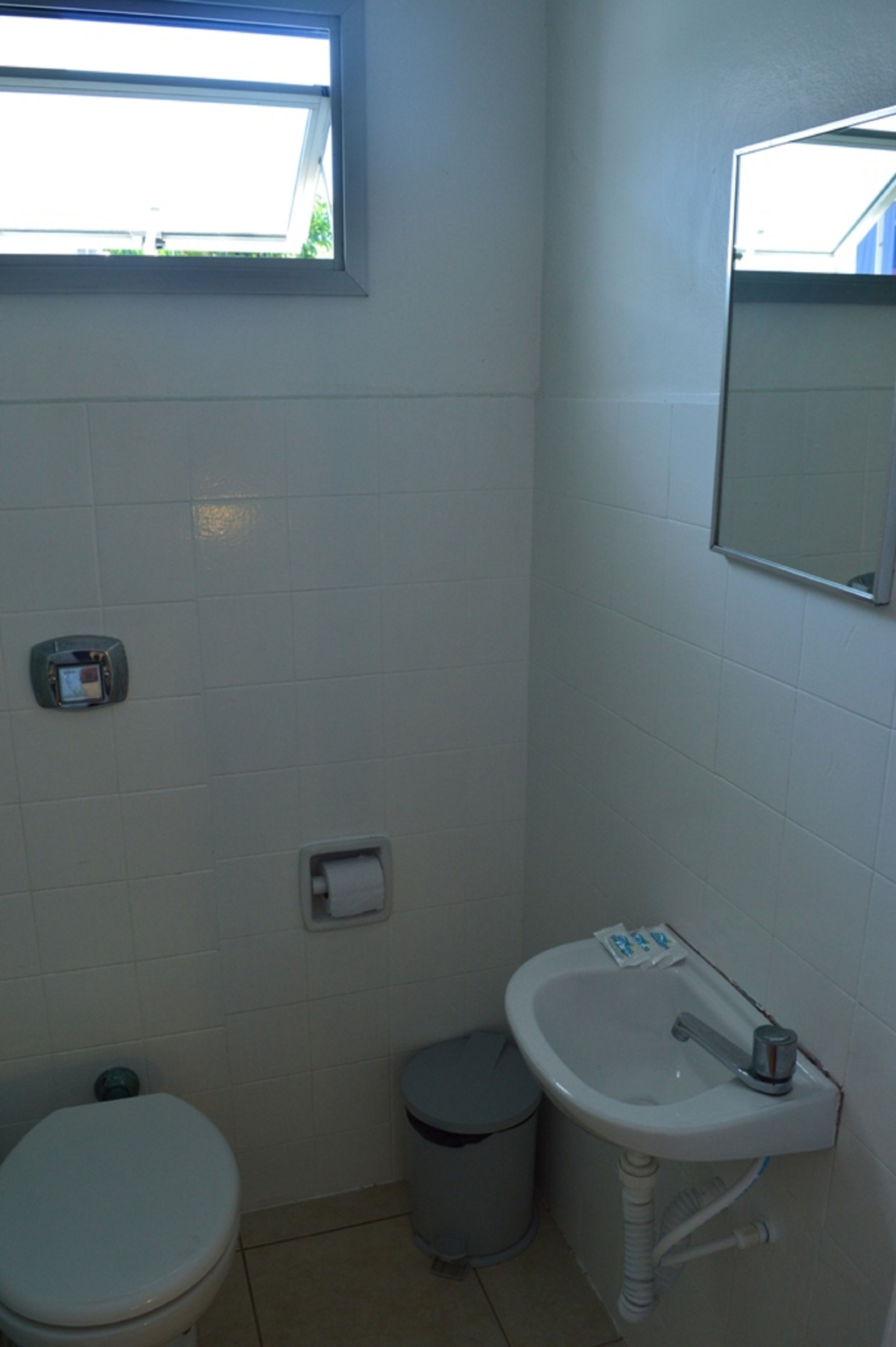Un baño pequeño con inodoro, lavabo y un cubo de basura. Hay una ventana sobre el inodoro y un espejo sobre el lavabo.