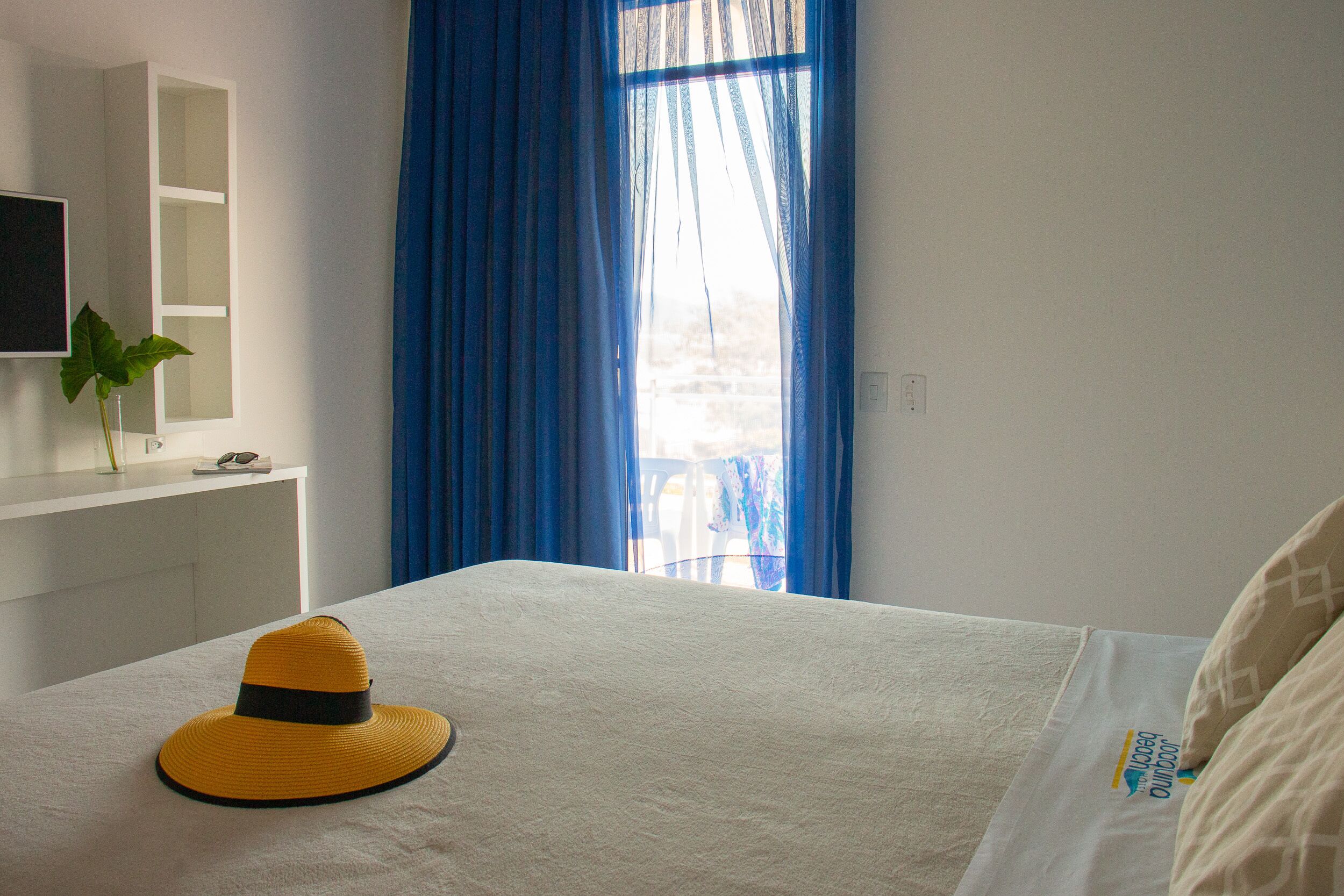 A hotel room with a bed, a TV on a white cabinet, and a window with blue curtains.