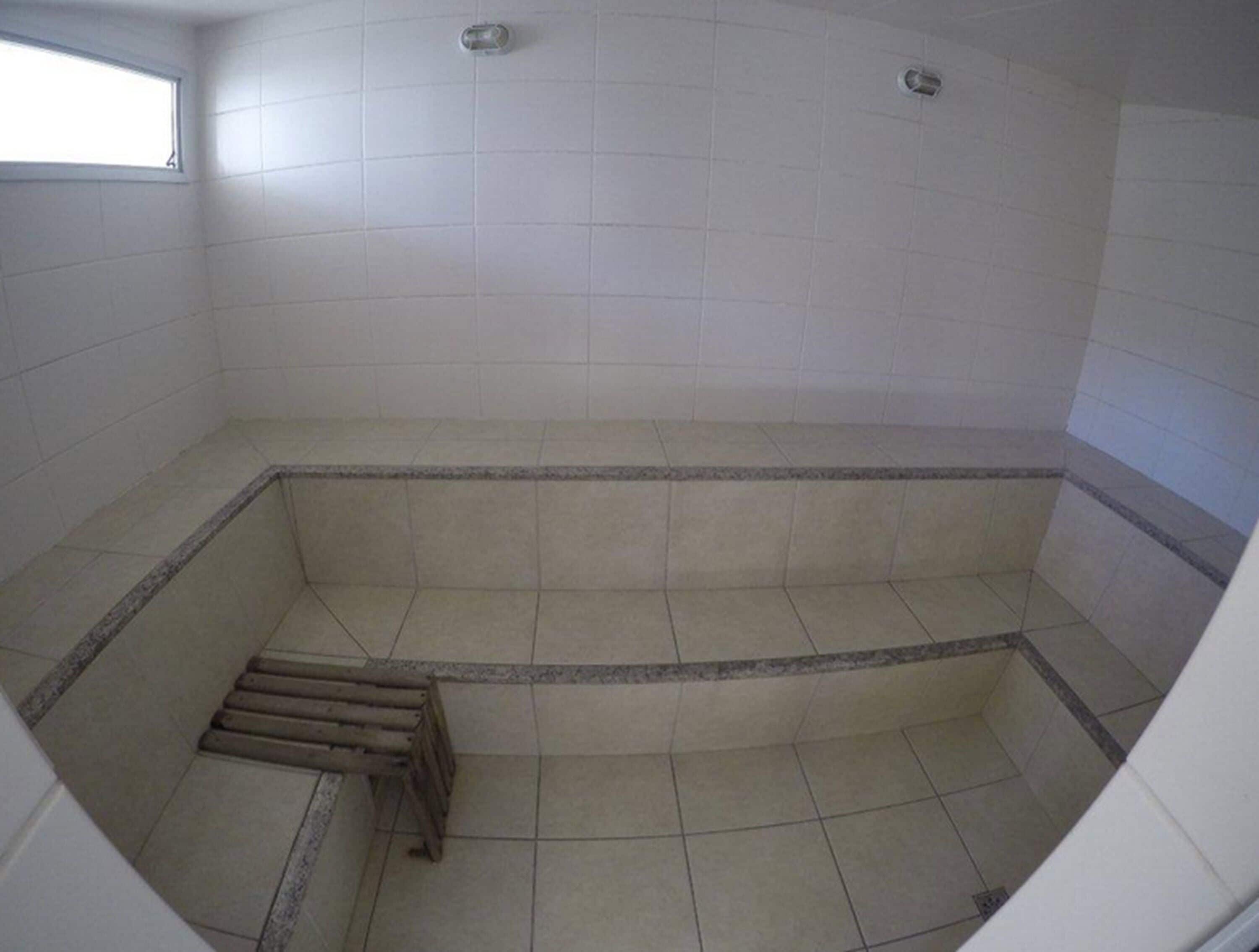 A small, empty room with white tiled walls and floor, a window on the left, and a shower area with a wooden bench.