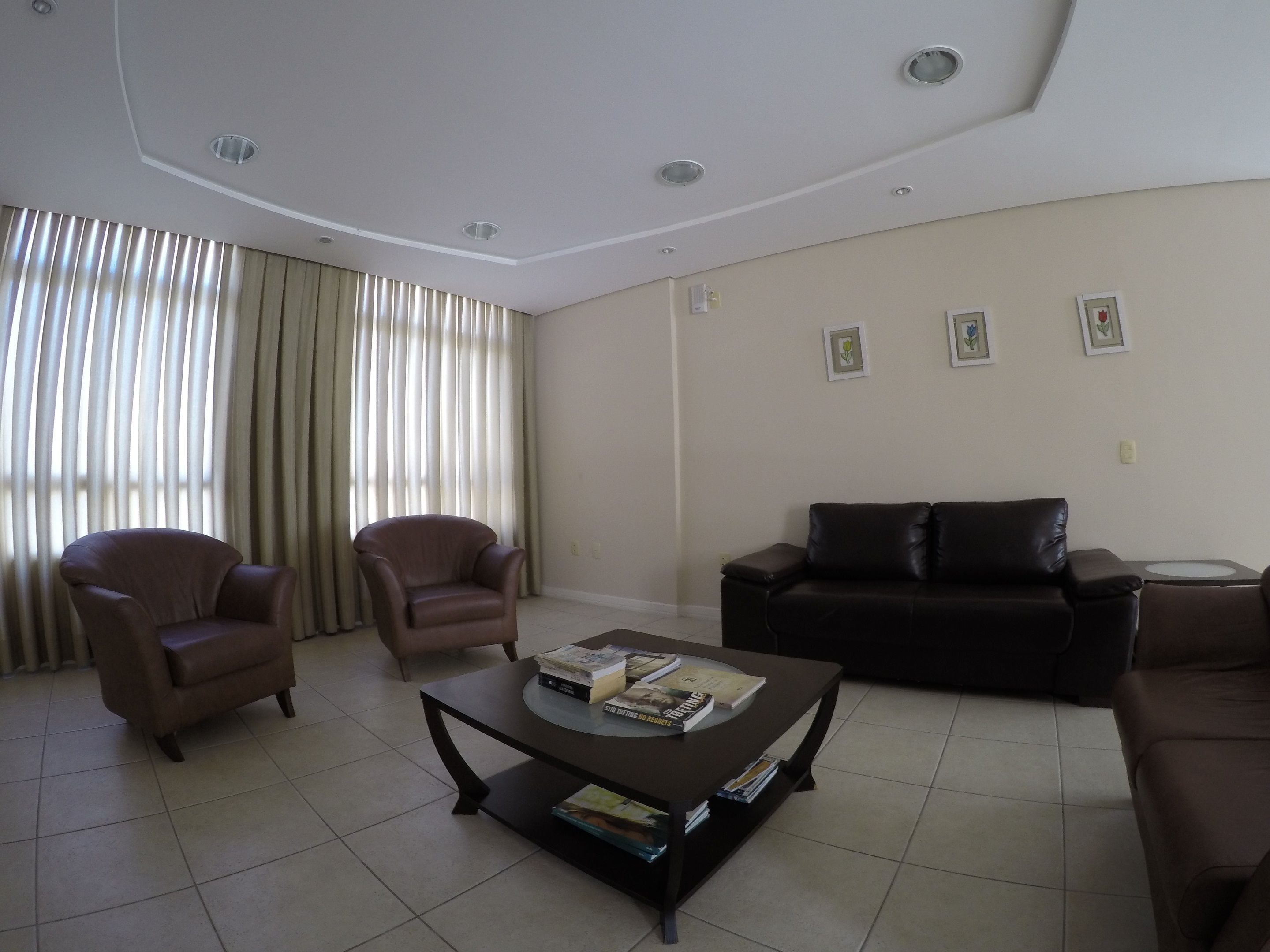 A living room with a brown leather sofa, two armchairs, a coffee table with books, and a large window with curtains.