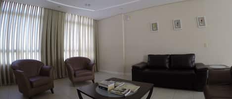 A living room with a brown leather sofa, two armchairs, a coffee table with books, and a large window with curtains.