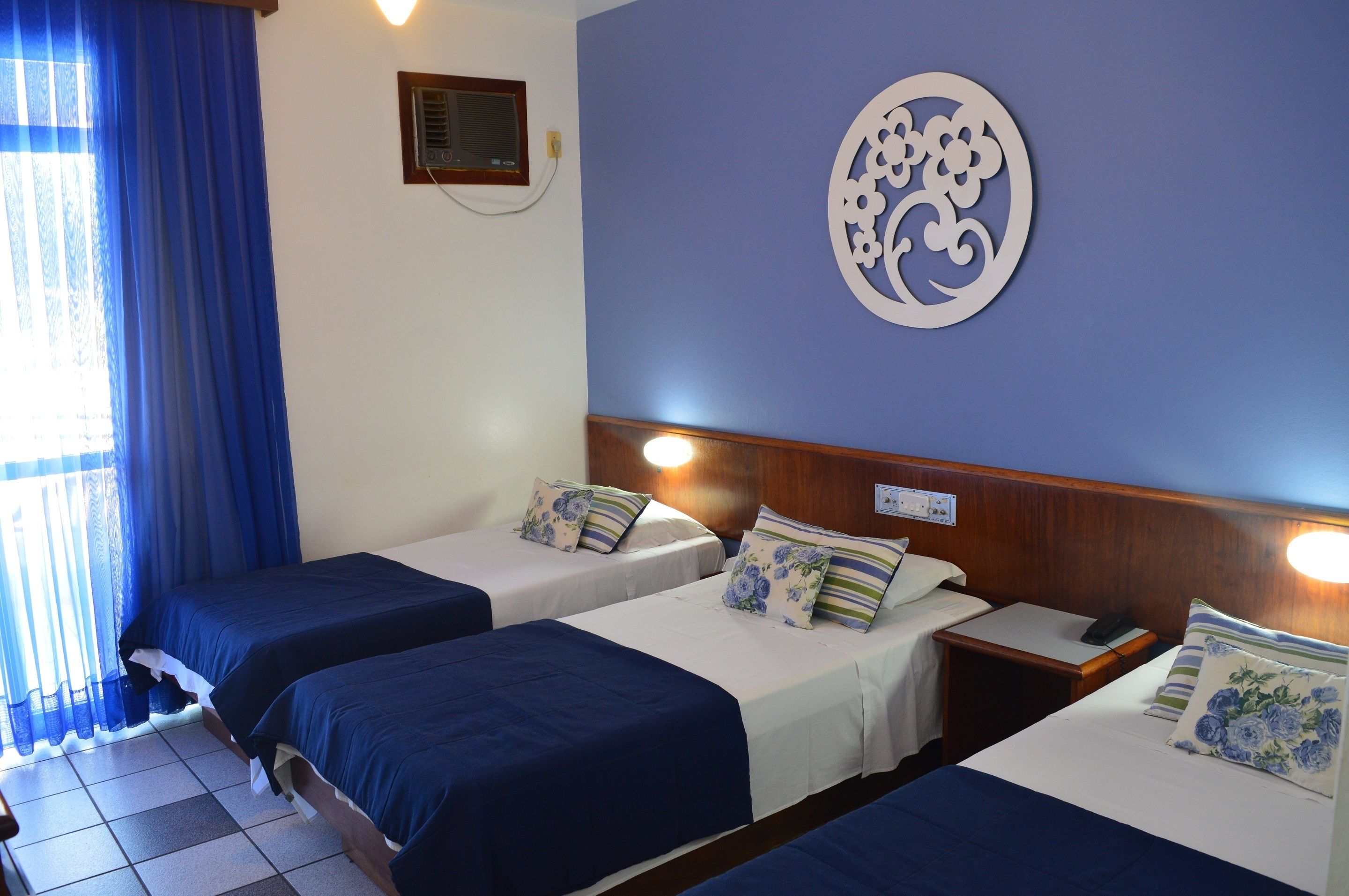 A hotel room with two single beds, a wooden headboard, a decorative wall piece, and a window with blue curtains.