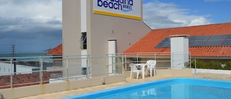 A swimming pool area with a view of the ocean, a building with a sign reading "Joaquina beach hotel", and a red-tiled roof.
