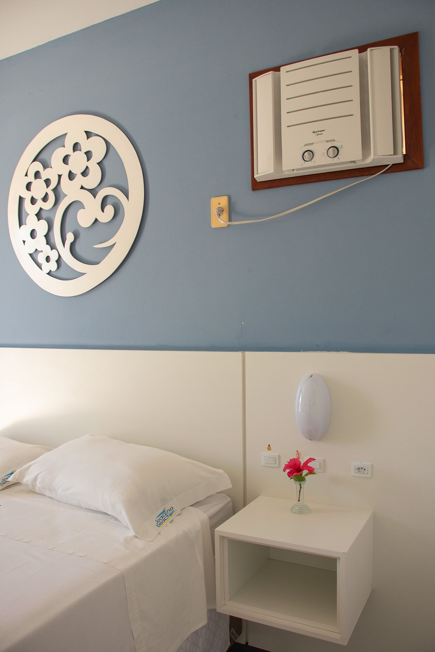 A hotel room with a bed, a bedside table with a vase of flowers, a decorative wall piece, and a thermostat on the wall.