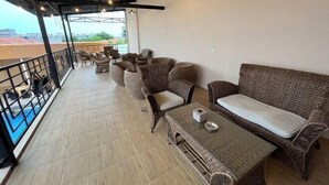 A covered outdoor seating area with wicker furniture, a table, and a view of the pool and buildings in the background. - Riez Palace Hotel (East Tegal)