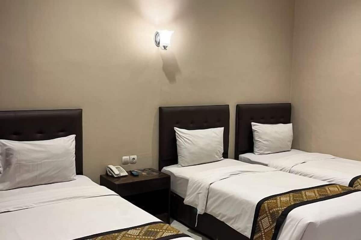 A hotel room with three beds, each with a headboard and white bedding. There is a nightstand with a phone and a wall-mounted light fixture.