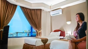 A hotel room with two beds, a large window with curtains, an air conditioning unit, and a person reading a book. - Grand Jambi (Jambi)