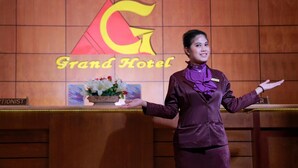 A hotel reception area with a Grand Hotel logo, a reception desk, and a floral arrangement. - Grand Jambi (Jambi)