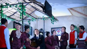 A group of people in uniform seated and standing in a room with a metal roof and green foliage decorations. - Grand Jambi (Jambi)