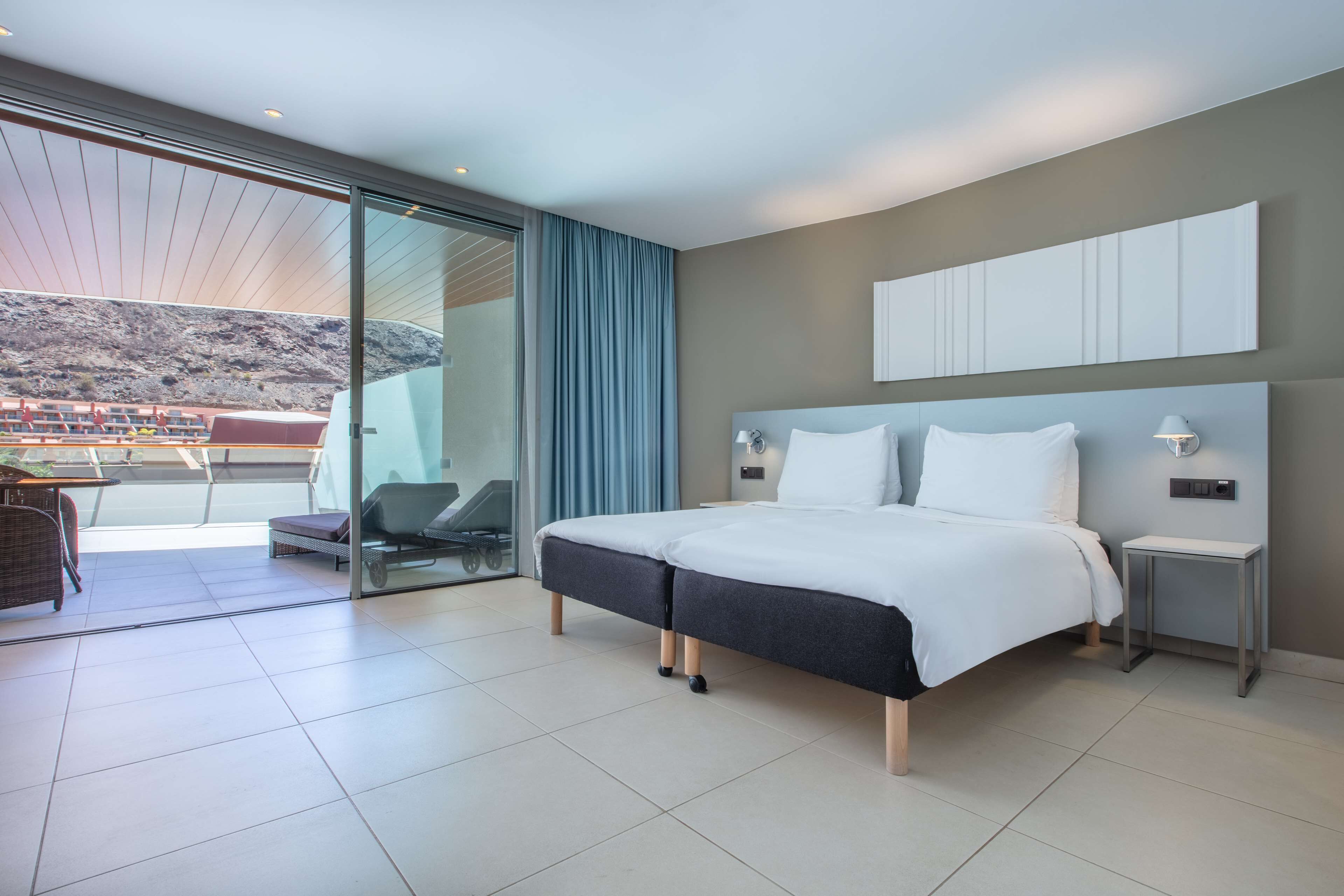 A modern hotel room with a large bed, a small bedside table, a chair, and a view of a mountainous landscape through a sliding glass door.