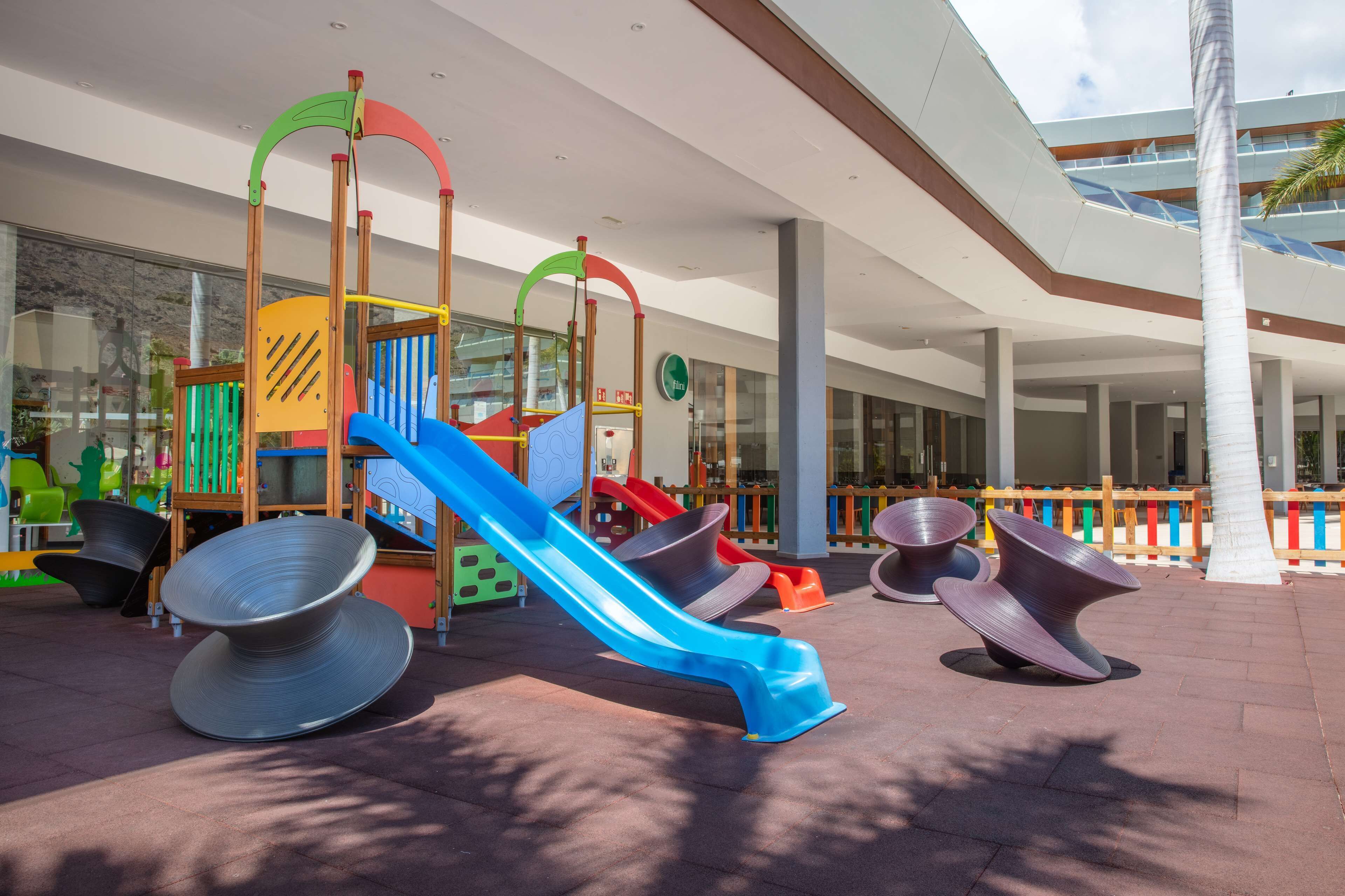 A children's play area with slides and climbing structures, outdoor seating, and a modern building facade.