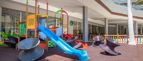 Zona de juegos infantiles con toboganes y estructuras para escalar, asientos al aire libre y fachada de edificio moderna.