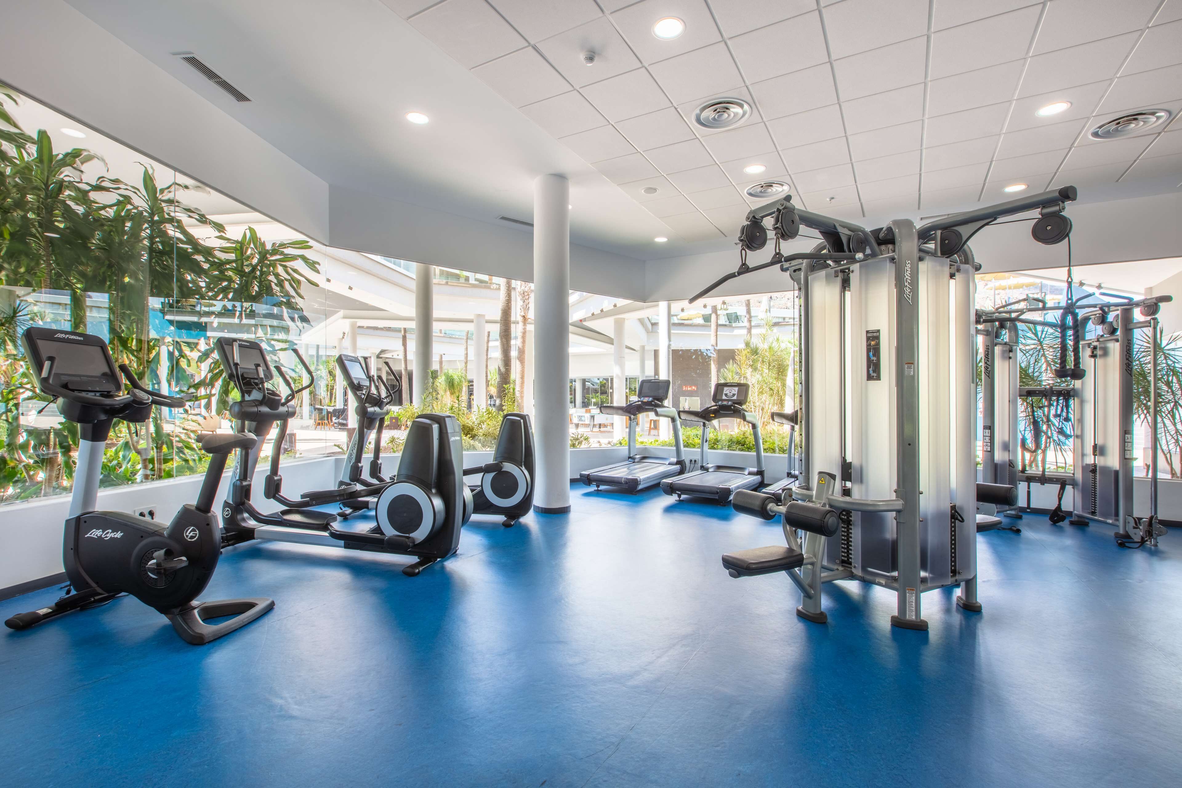 A modern gym with various exercise equipment, large windows, and a blue floor.