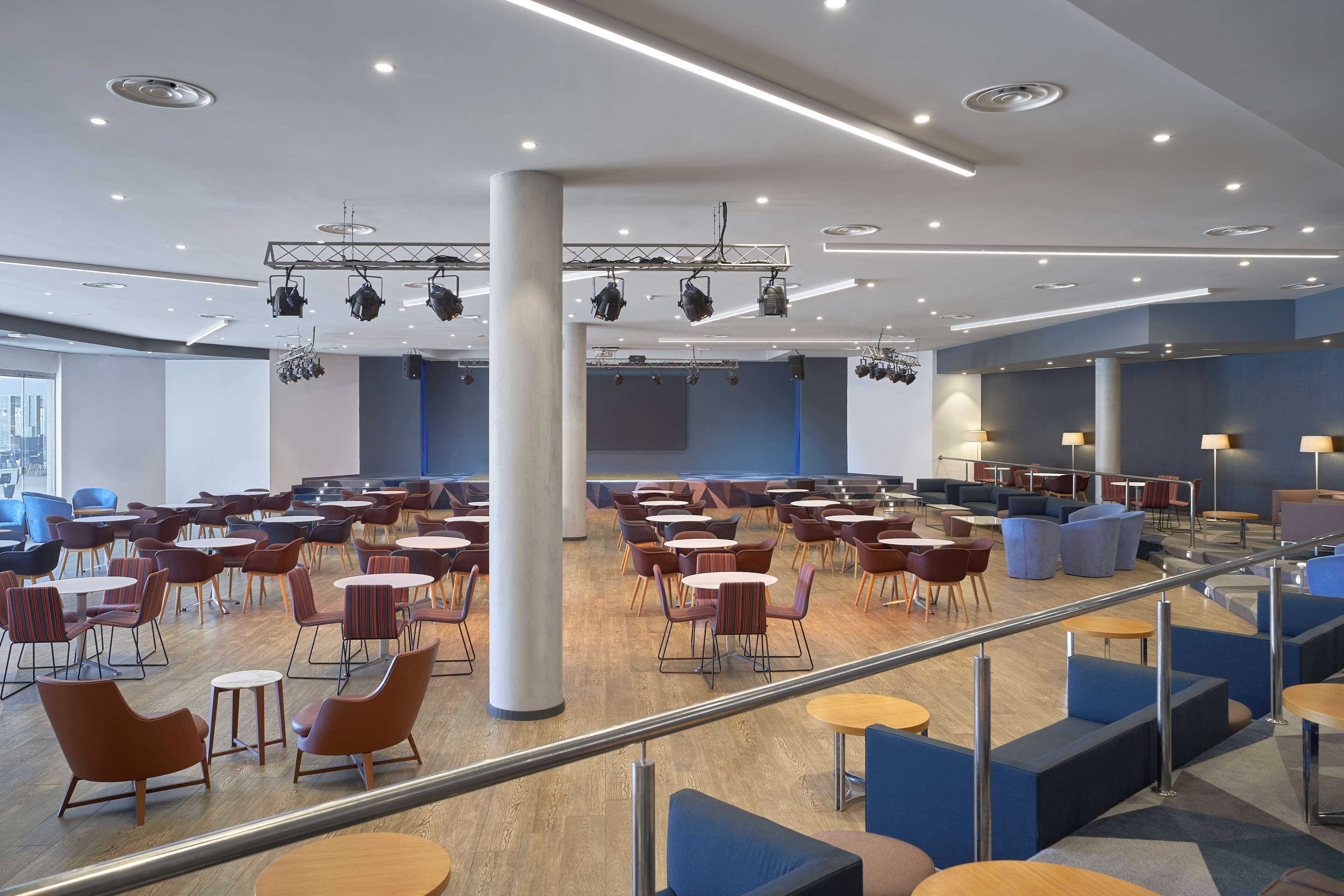 Amplio espacio interior con diversas disposiciones de asientos, incluidas mesas y sillas, y un escenario con equipo de iluminación.